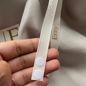 May include: Close-up of a cream-colored ribbon with the word "Eray" printed on it. Two white circular hook-and-loop fasteners are attached to the ribbon. The background is a light gray fabric.