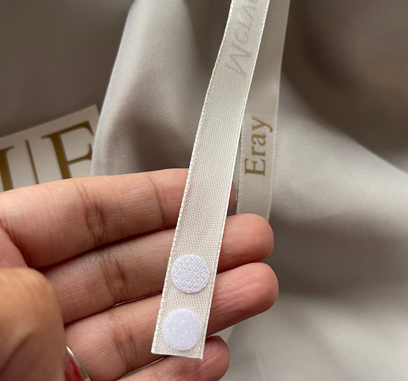 May include: Close-up of a cream-colored ribbon with the word "Eray" printed on it. Two white circular hook-and-loop fasteners are attached to the ribbon. The background is a light gray fabric.