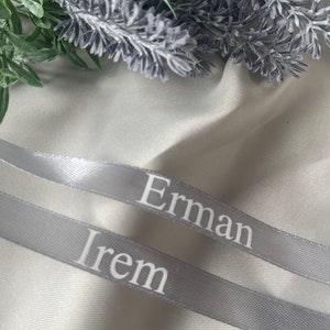 May include: Two gray satin ribbons with the names "Erman" and "Irem" printed in white. The ribbons are positioned diagonally across a white fabric, with green and purple floral accents in the background.