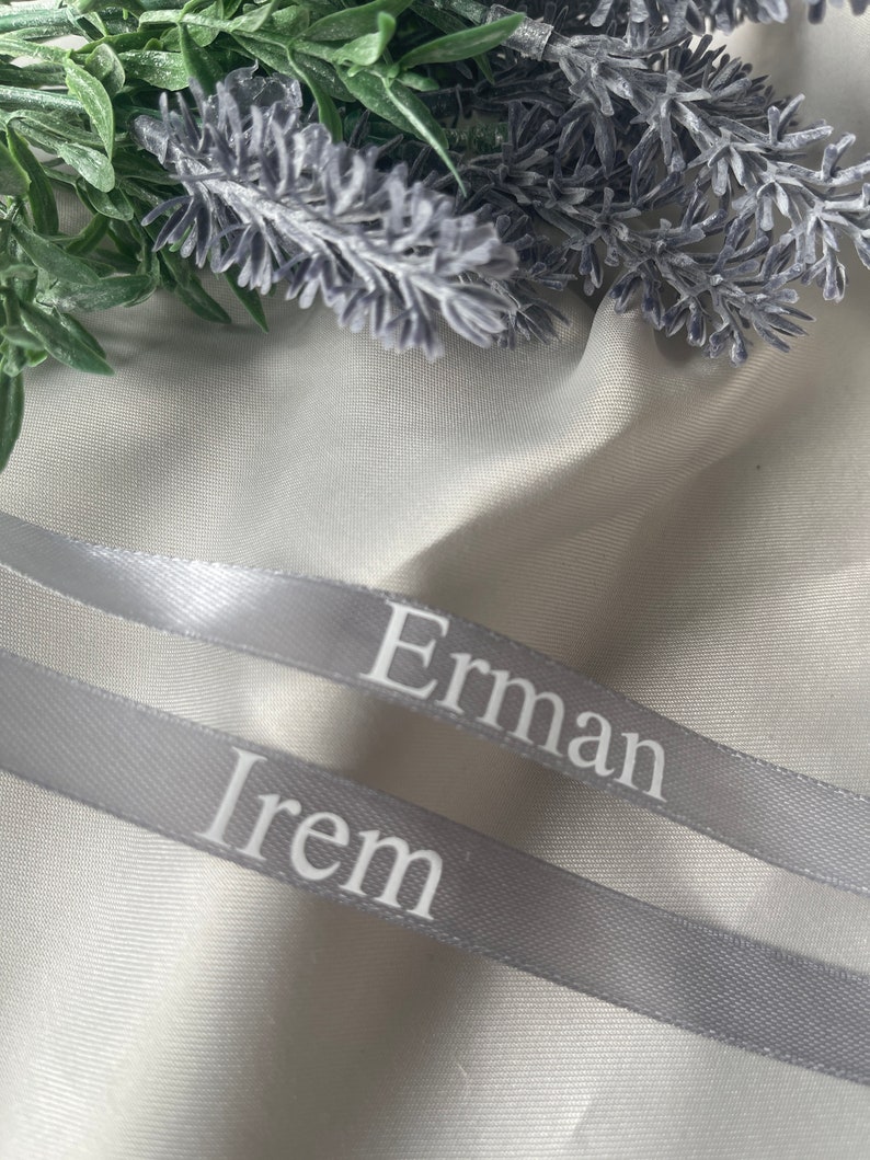 May include: Two gray satin ribbons with the names "Erman" and "Irem" printed in white. The ribbons are positioned diagonally across a white fabric, with green and purple floral accents in the background.