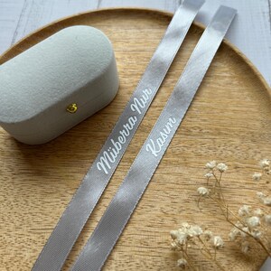 May include: A light gray velvet ring box with a gold clasp sits on a wooden tray, accompanied by two gray ribbons. The ribbons are printed with the names "M&uuml;berra Nur" and "Kasım" in white script.