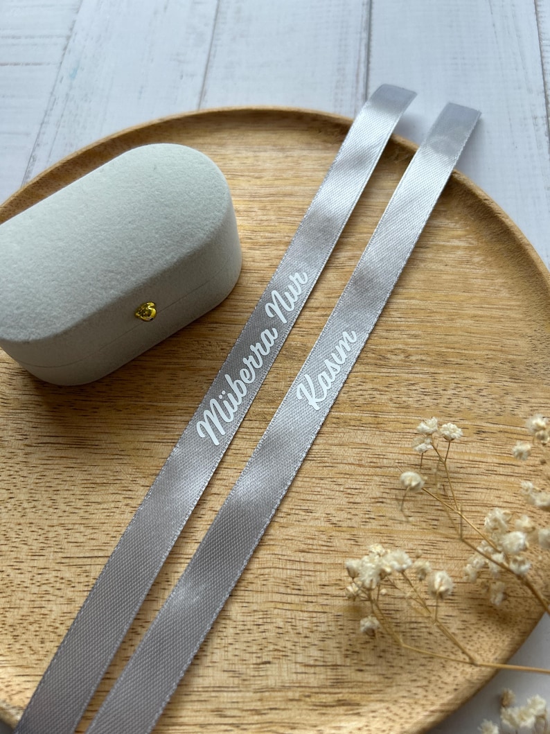 May include: A light gray velvet ring box with a gold clasp sits on a wooden tray, accompanied by two gray ribbons. The ribbons are printed with the names "M&uuml;berra Nur" and "Kasım" in white script.