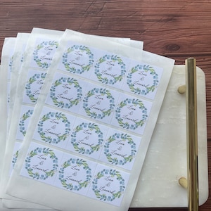 May include: A stack of white square stickers with a floral wreath design in blue and green. The text "Lena & Lennard" is centered within the wreath. The stickers are on a white marble tray with gold handles.