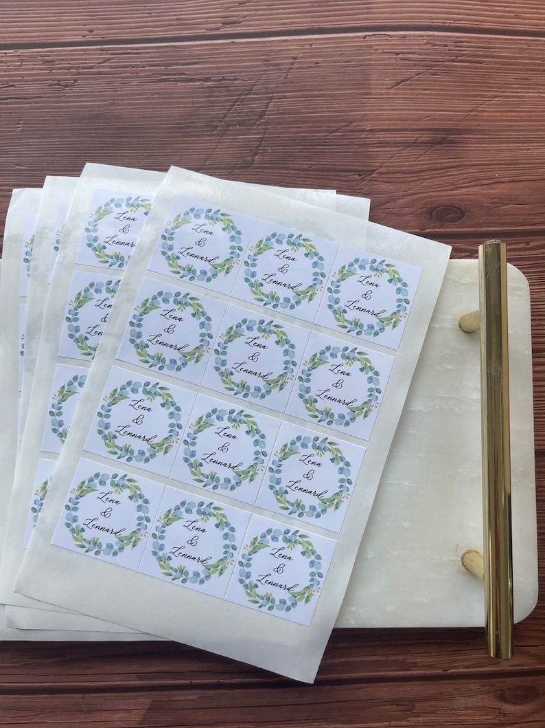 May include: A stack of white square stickers with a floral wreath design in blue and green. The text "Lena & Lennard" is centered within the wreath. The stickers are on a white marble tray with gold handles.