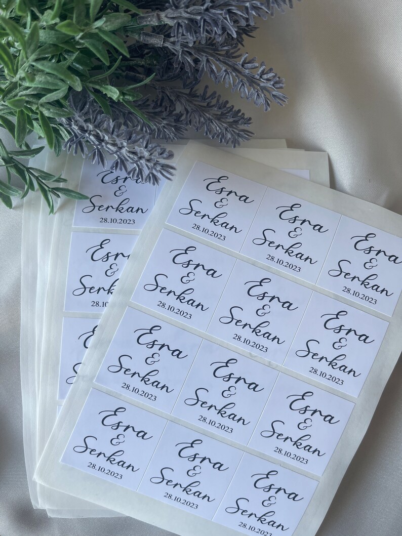 May include: White square stickers with black calligraphy text that reads "Esra & Serkan 28.10.2023". The stickers are stacked on top of each other. The background includes a sprig of lavender.