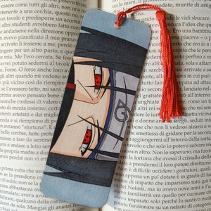 Può includere: Segnalibro con illustrazione di un personaggio in stile anime, con fascia nera, occhi rossi e nappa rossa. Il segnalibro è posto sulle pagine di un libro aperto.