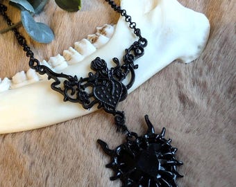 May include: Black metal necklace with a heart and sunburst pendant. The necklace features a delicate chain and ornate floral and heart-shaped details. The jewelry is displayed on a bone against a brown fur-like background.