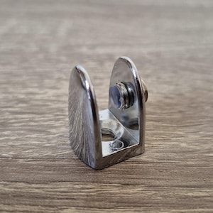 Op de afbeelding: Een glanzende, verchroomde glazen plankbeugel. De beugel heeft een U-vormig ontwerp met een schroef voor het bevestigen van het glas. De beugel is ontworpen om een glazen plank te ondersteunen en is gemaakt van metaal.
