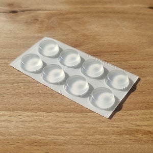 May include: A sheet of clear adhesive pads, arranged in a rectangular pattern on a wooden surface. The pads are designed to protect surfaces from scratches and damage.