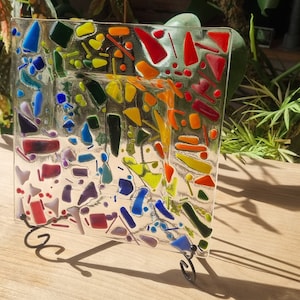 Può includere: Un piatto quadrato di vetro con un design a mosaico di pezzi di vetro colorati. Il piatto è su un supporto e ha uno schema di colori arcobaleno.