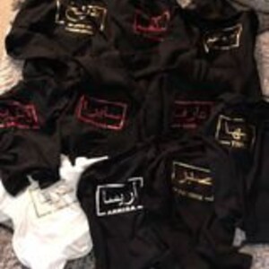Puede incluir: Una pila de sudaderas con capucha negras con un logotipo bordado en dorado, rojo y blanco que dice "America" en la parte delantera. Las sudaderas con capucha est&aacute;n dobladas y apiladas unas sobre otras. Hay una camiseta blanca debajo de las sudaderas con capucha.