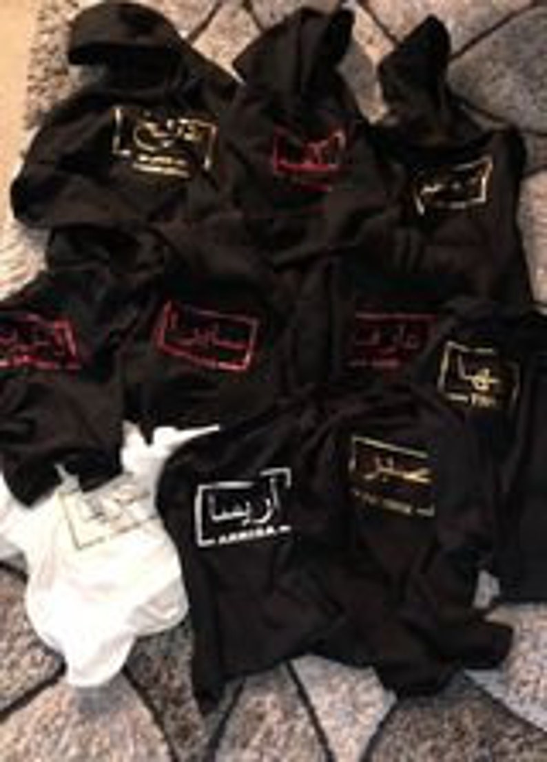 Puede incluir: Una pila de sudaderas con capucha negras con un logotipo bordado en dorado, rojo y blanco que dice "America" en la parte delantera. Las sudaderas con capucha est&aacute;n dobladas y apiladas unas sobre otras. Hay una camiseta blanca debajo de las sudaderas con capucha.
