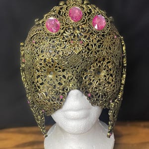 May include: Ornate, antique-style mask in gold-tone metal, featuring intricate filigree patterns. The mask is adorned with three large, pink gemstones across the top and smaller pink accents. It is displayed on a white mannequin head, suggesting costume or decorative use.