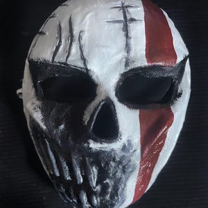 Può includere: Una maschera a teschio bianca con orbite nere e una mascella nera. Una striscia rossa verticale corre lungo il lato destro. La maschera ha segni neri ed è probabilmente per costumi o esposizioni.