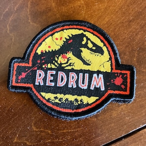 Puede incluir: Un parche bordado negro, amarillo y rojo con una silueta de dinosaurio y la palabra "REDRUM" en letras blancas.