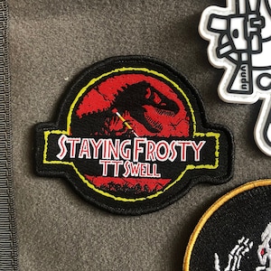 May include: Black and yellow embroidered patch with a red dinosaur silhouette inside a yellow circle. The text "STAYING FROSTY TTSWELL" is written in white on a black banner.