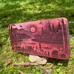 Puede incluir: Una placa de madera con una imagen grabada con láser de una puesta de sol sobre un río y montañas. La placa está hecha de madera marrón rojiza y tiene un acabado natural.