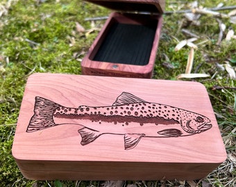 Puede incluir: Una caja de madera con tapa abatible, que presenta una imagen grabada de una trucha. La caja está hecha de madera de cedro y tiene un color marrón rojizo natural. La tapa está abierta, revelando un interior negro.