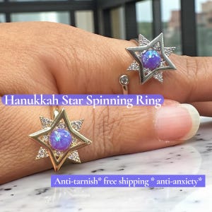 Può includere: Primo piano di due anelli con design a stella. Gli anelli presentano un'incastonatura a forma di stella con una pietra blu-viola al centro, circondata da pietre chiare più piccole. Il testo "Hanukkah Star Spinning Ring" è visibile.