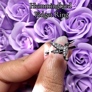 May include: A silver ring with a hummingbird design, featuring clear rhinestones. The ring is on a finger against a background of purple roses. The text "Hummingbird Fidget Ring" is displayed above the ring.