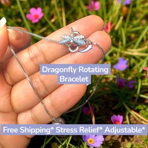 May include: A silver-colored bracelet with a dragonfly design. The dragonfly has blue accents and is connected to a delicate chain. The text "Dragonfly Rotating Bracelet" is visible.