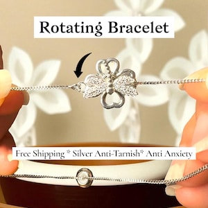 May include: A silver rotating bracelet with a clover-shaped charm, adorned with sparkling accents. The bracelet is held between two hands, showcasing its delicate chain design. The text "Rotating Bracelet" is displayed above the bracelet.