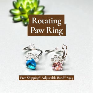 May include: Two silver-toned rotating paw rings with a blue and pink gemstone. The rings have a paw-shaped design with clear accent stones. The image includes the text "Rotating Paw Ring".