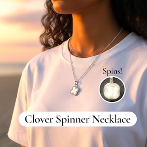 Op de afbeelding: Een zilveren klaver spinner ketting met een witte klaver hanger, getoond op een persoon die een wit t-shirt draagt. De ketting heeft een kleine, ronde, draaiende hanger. De tekst op de afbeelding luidt "Spins!" en "Clover Spinner Necklace."