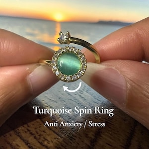 Könnte beinhalten: Ein goldfarbener Ring mit einem türkis farbenen Mittelstein, umgeben von kleinen, klaren Steinen. Der Ring hat eine Drehfunktion. Der Text "Turquoise Spin Ring" und "Anti Anxiety / Stress" ist sichtbar.