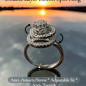 May include: A silver-toned double layer zircon spin ring featuring a central cluster of clear stones and a rotating band. The text reads Double Layer Zircon Spin Ring and Anti-Anxiety/Stress * Adjustable fit * Anti-Tarnish with curved arrows.