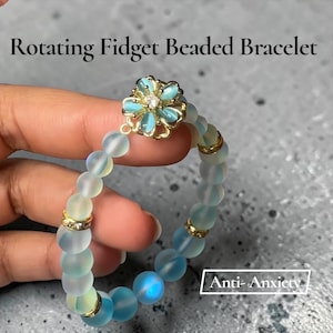 May include: A beaded bracelet featuring translucent light blue frosted beads, gold-toned spacers, and a central flower-shaped charm with a clear stone. The text "Rotating Fidget Beaded Bracelet" and "Anti-Anxiety" are visible in the image.