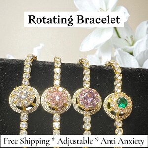 Könnte beinhalten: Vier goldfarbene Dreharmbänder mit unterschiedlich farbigen Mittelsteinen: klar, rosa, rosa und grün. Die Armbänder sind mit kleinen, klaren Steinen besetzt. Der Text "Rotating Bracelet" steht oben.