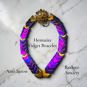 May include: A hematite fidget bracelet with a purple and gold color scheme. The bracelet has a flower-shaped clasp with a pearl in the center. The text on the image says "Hematite Fidget Bracelet", "Anti-Stress", and "Reduce Anxiety".