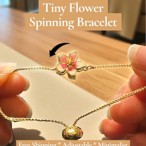 May include: A gold-colored bracelet with a tiny, spinning flower charm. The flower has pink and white petals with a gold center. The bracelet is held between two hands. Text on the image reads "Tiny Flower Spinning Bracelet".