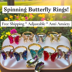 May include: A collection of gold-toned, adjustable rings with colorful, spinning butterfly designs. The butterflies feature various colors, including pink, red, blue, and green, and are displayed on a white surface. The text "Spinning Butterfly Rings!" is visible.