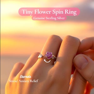Könnte beinhalten: Ein Tiny Flower Spin Ring aus Sterlingsilber mit einem rosa Blumenmuster. Der Ring wird am Finger einer Person gezeigt, wobei sich das Blumenelement dreht. Der Text "Stress/ Anxiety Relief" ist ebenfalls sichtbar.