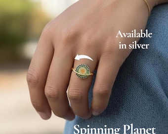 May include: A gold-colored ring and bracelet set with a planet design. The ring is on a finger, and the bracelet is on the wrist. The planet design features green, orange, and blue accents. The text "Spinning Planet Ring & Bracelet" is visible.
