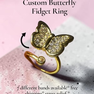 Könnte beinhalten: Ein goldfarbener Fidget-Ring mit einem Schmetterlingsmotiv, das mit klaren Steinen besetzt ist. Der Ring hat ein offenes Design mit einem kleinen Stein an einem Ende. Der Text auf dem Bild lautet: Custom Butterfly Fidget Ring, 7 different bands available.