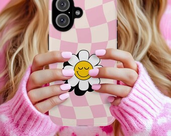 Puede incluir: Una funda de teléfono con un patrón de cuadros rosa y blanco y un gráfico de margarita. La margarita tiene pétalos blancos, un centro negro y una cara sonriente amarilla. La funda del teléfono la sostiene una persona que lleva un suéter rosa.