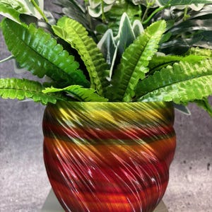 May include: A decorative plant pot with a swirling design in shades of red, orange, and yellow. The pot is filled with a variety of green plants, including ferns and ivy. The pot is sitting on a white surface.