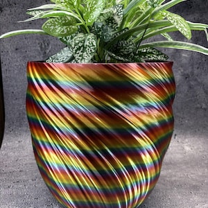 May include: A plant pot with a spiralling, ribbed texture and a metallic rainbow gradient finish containing a green and white variegated houseplant. The pot features a continuous, swirling pattern that shifts through colours of red, orange, yellow, green, blue, and purple.
