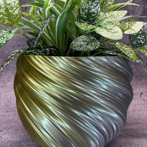 Puede incluir: Una maceta iridiscente verde y plateada con un patrón de remolino. Una planta verde con variegación blanca está creciendo en la maceta.