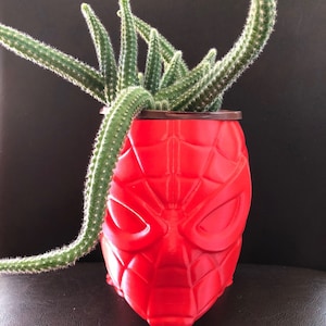 Puede incluir: Una maceta de plástico roja con forma de cabeza de Spider-Man con una planta de cactus verde que crece en la parte superior.