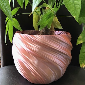 May include: A decorative, swirled planter in shades of pink, brown, and white holds a green plant with large leaves. The planter has a unique, textured design and sits on a dark surface.