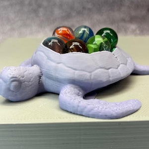 Könnte beinhalten: Ein lavendelfarbener, schildkrötenförmiger Behälter mit mehreren bunten Murmeln. Die Murmeln sind in verschiedenen Farben gehalten, darunter Orange, Grün, Blau und Braun. Der Behälter steht auf einer hellen Oberfläche.
