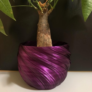 May include: A vibrant purple, metallic-looking planter pot cradles a small tree. The pot has a unique, swirled design, and the tree features green leaves and a brown trunk. The background is a dark color, and the pot sits on a white surface.