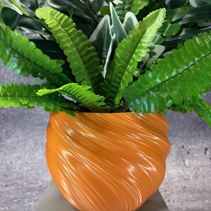 May include: An orange, swirled-design planter filled with a variety of green artificial plants. The planter has a glossy finish and sits on a light gray surface. The plants include ferns and other leafy varieties.