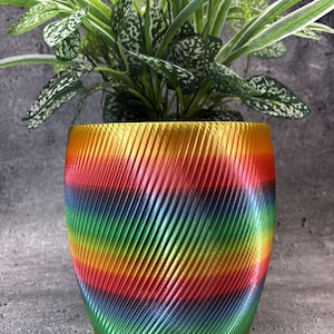 Peut inclure: Un pot de fleurs cylindrique présentant un dégradé arc-en-ciel vibrant de jaune, orange, rouge, bleu et vert. La surface extérieure est texturée avec un motif répétitif de crêtes diagonales en relief créant un effet dynamique.