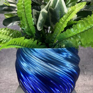 May include: A vibrant blue planter with a distinct swirling, ribbed texture containing a lush arrangement of artificial green foliage. The plant features various leaf shapes, including serrated fern-like fronds and broad, variegated leaves with white edges.