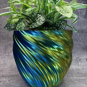 Puede incluir: Maceta decorativa azul y verde iridiscente con un diseño en espiral. Contiene una planta verde con hojas moteadas de blanco. La superficie texturizada y el degradado de color de la maceta contrastan con el aspecto natural de la planta. Un artículo de decoración del hogar.